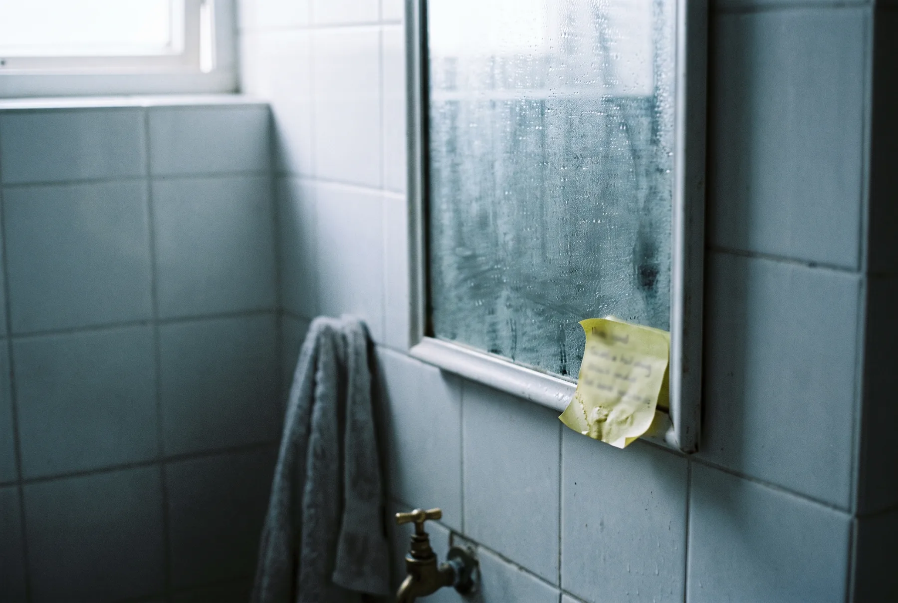 Een beslagen badkamerspiegel in koel ochtendlicht met een geel briefje aan de onderrand geplakt, er is niets in de spiegel te zien
