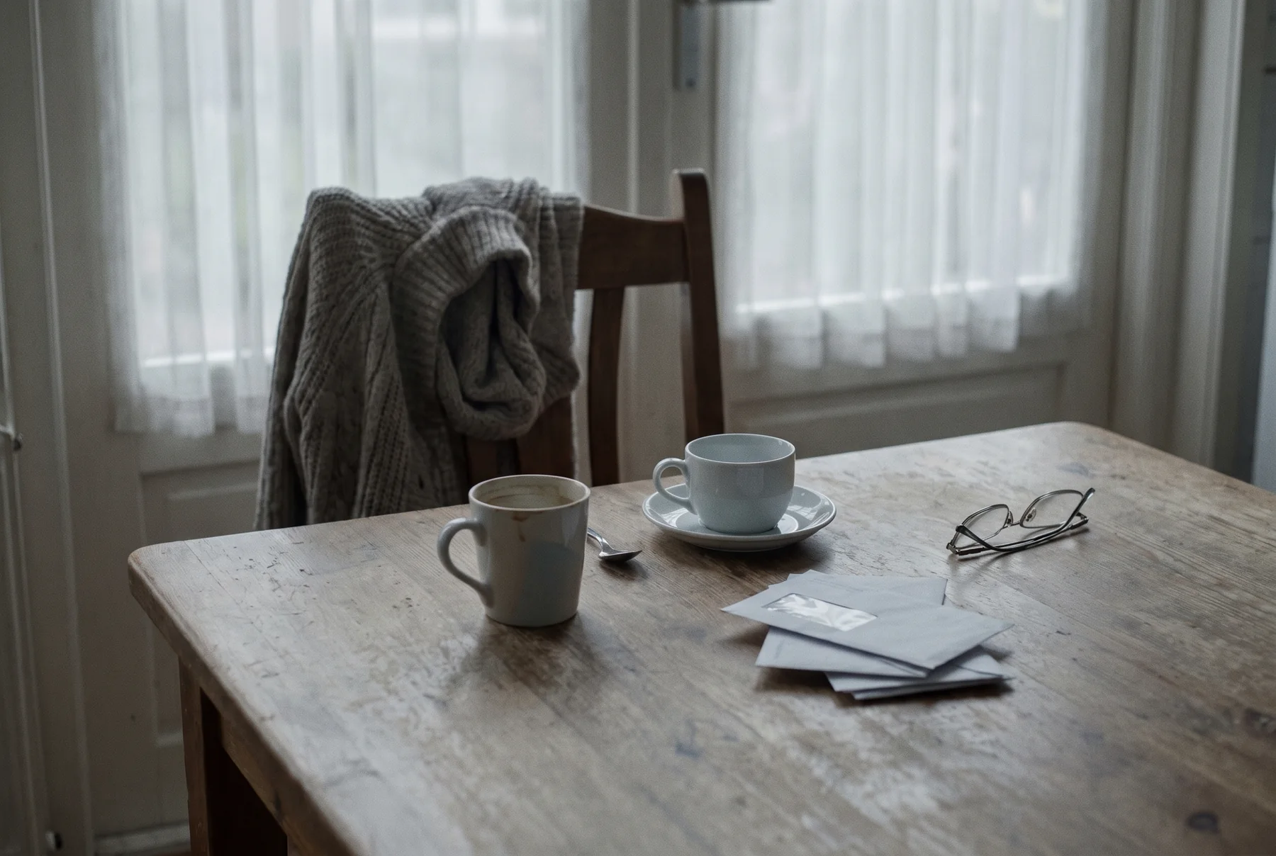 Een keukentafel met een stoel naar achteren geschoven, een vest over de rugleuning en twee kopjes waarvan er een onaangeroerd is