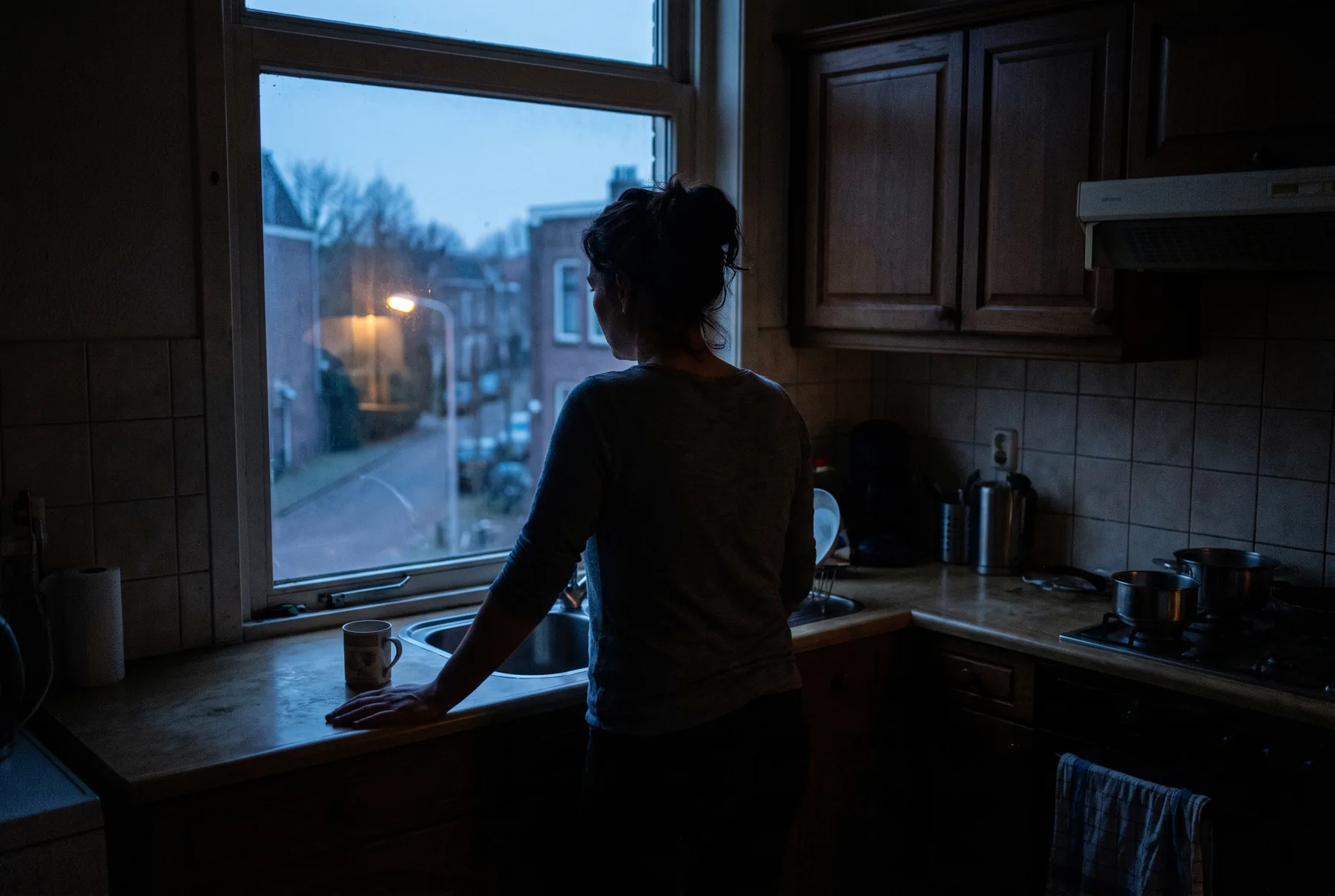 Een vrouw alleen in een donkere keuken bij het raam in de schemering, hand plat op het aanrecht, buiten een straat met een brandende lantaarn