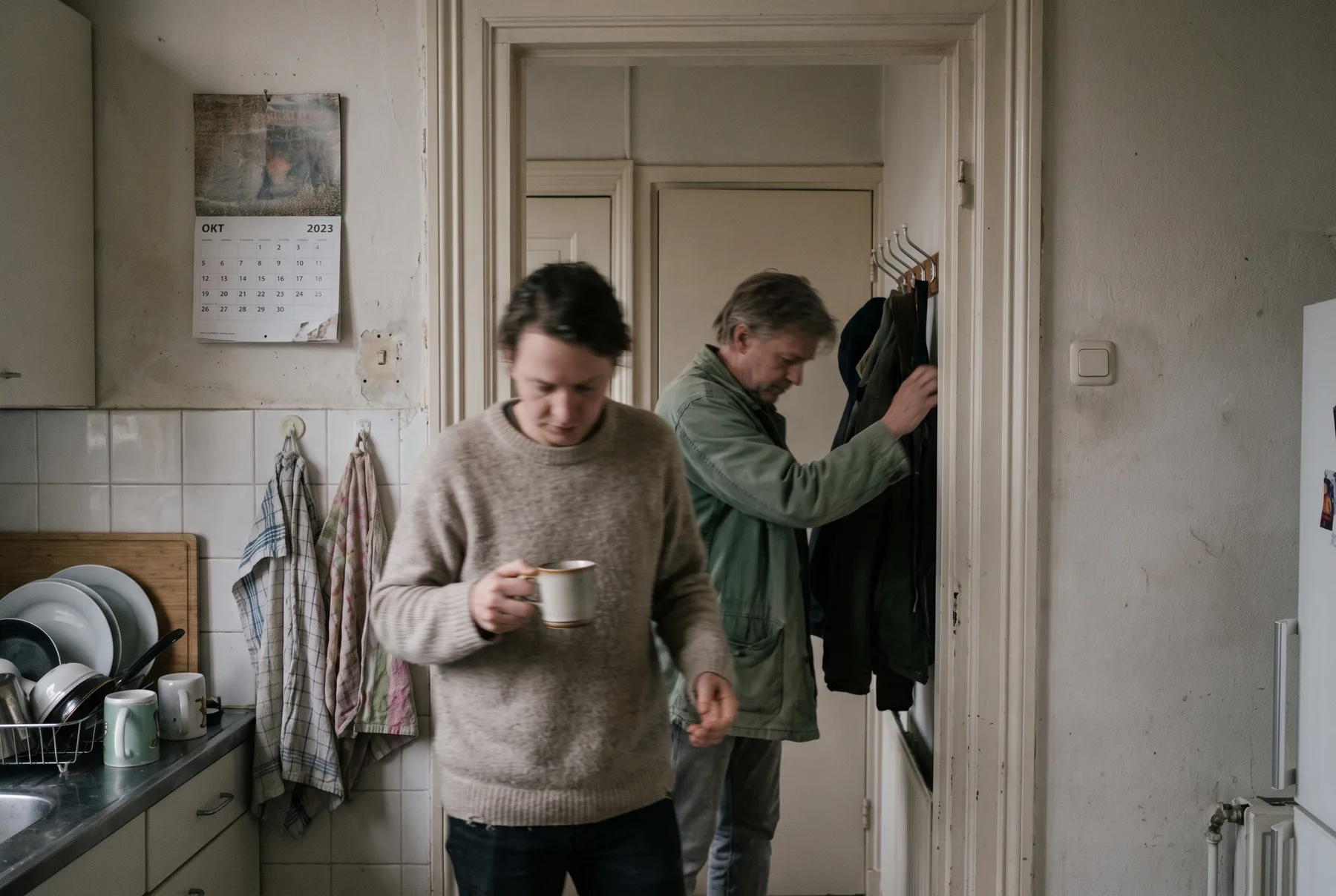 Twee mensen passeren elkaar in een smalle keukendeur, de een met een kop koffie, de ander reikt naar een jas, geen van beiden kijkt op