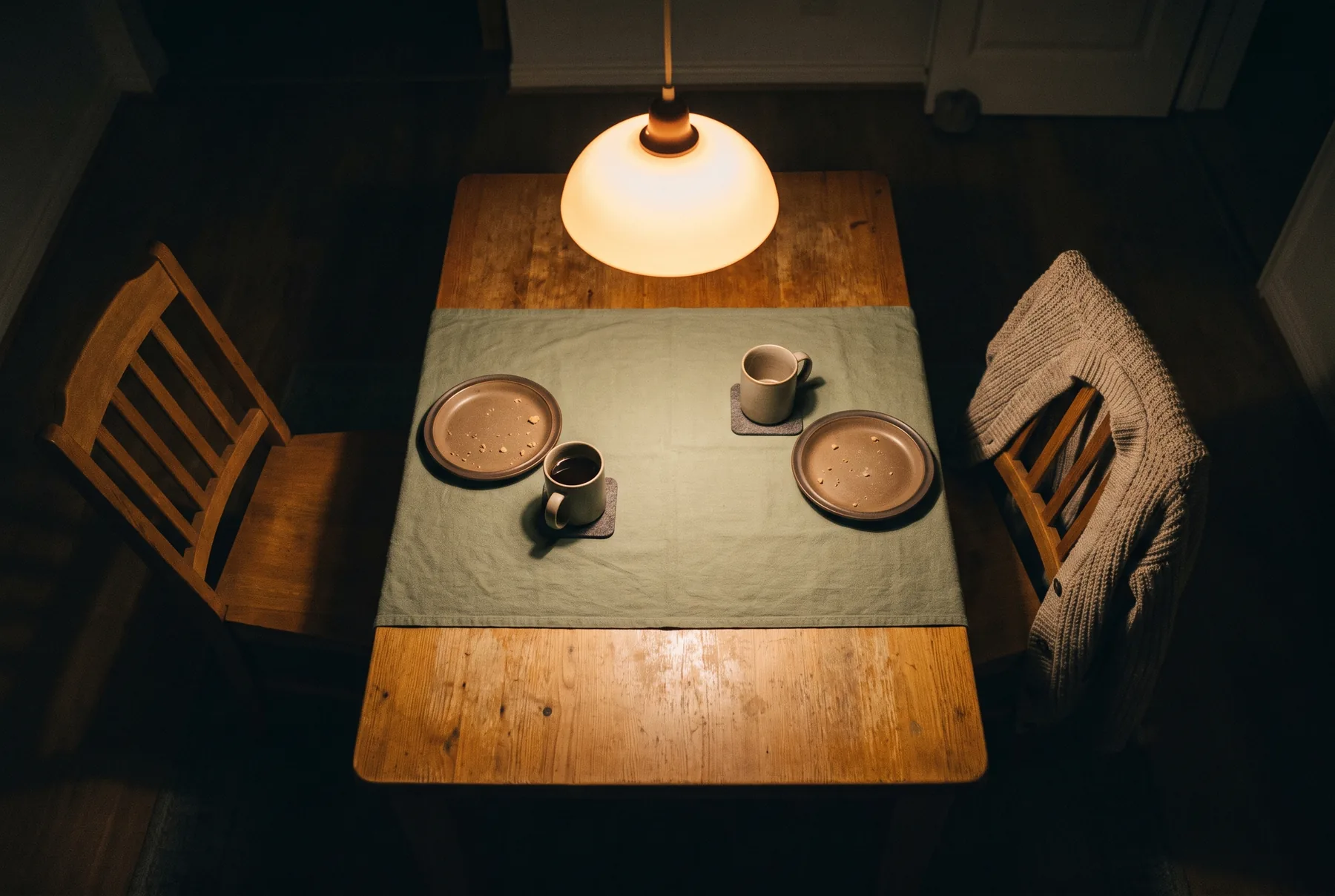 Een eettafel van bovenaf in de avond, twee plaatsen tegenover elkaar, een stoel achteruitgeschoven en leeg en op de andere een vest