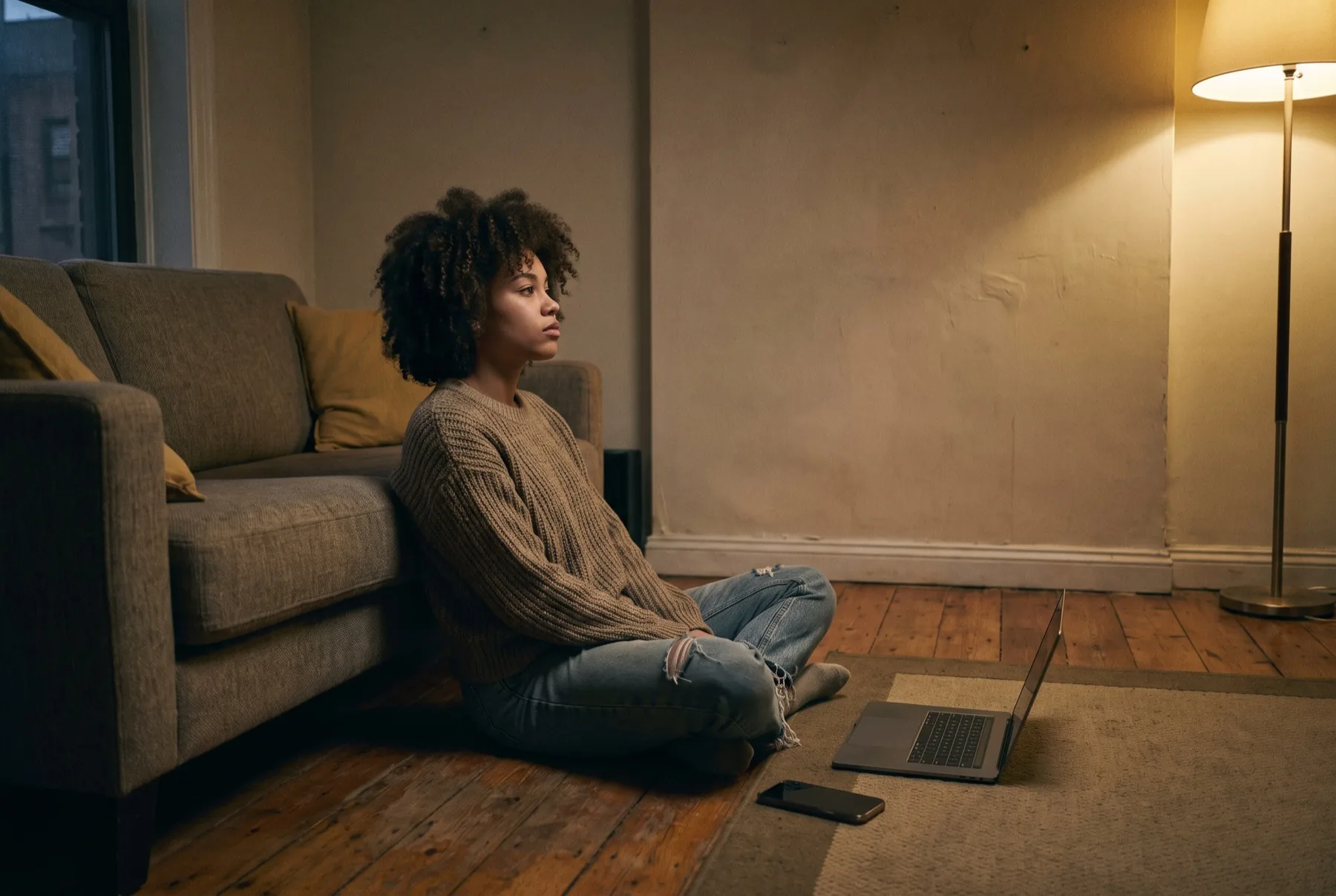 Een jonge vrouw op de vloer tegen de bank, laptop dicht, telefoon omgekeerd